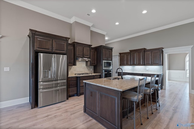 kitchen featuring crown molding, light hardwood / wood-style flooring, appliances with stainless steel finishes, light stone countertops, and a center island with sink