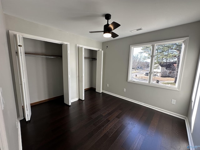 unfurnished bedroom featuring two closets, visible vents, dark wood-type flooring, ceiling fan, and baseboards