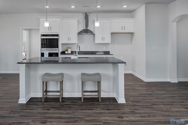 kitchen featuring dark wood-type flooring, stainless steel double oven, white cabinets, a kitchen island with sink, and wall chimney range hood