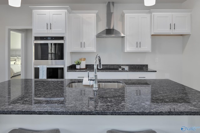 kitchen featuring white cabinets, stainless steel double oven, dark stone counters, and wall chimney exhaust hood