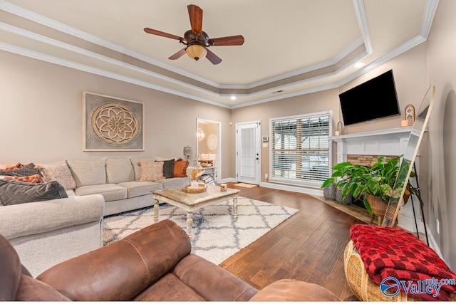 living room with a ceiling fan, a tray ceiling, a fireplace, ornamental molding, and hardwood / wood-style flooring