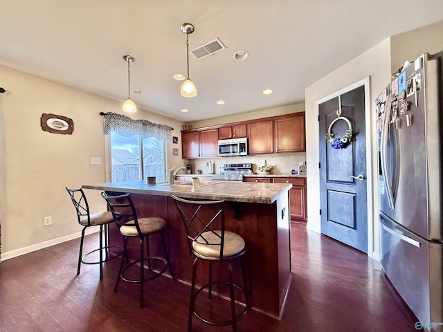 kitchen featuring appliances with stainless steel finishes, an island with sink, decorative backsplash, hanging light fixtures, and dark wood-type flooring