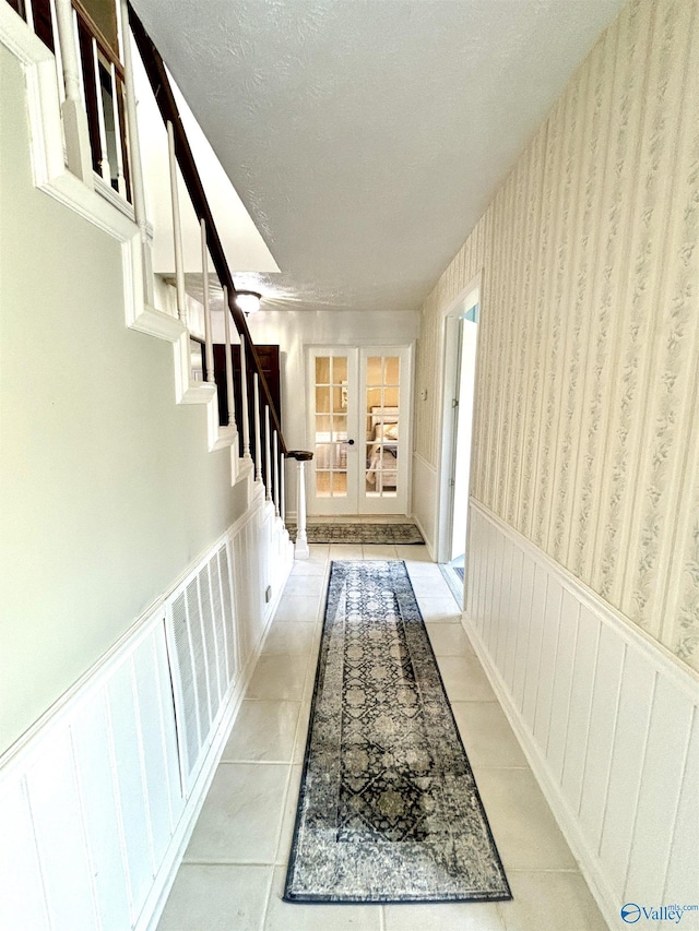 corridor featuring a wainscoted wall, a textured ceiling, wallpapered walls, stairway, and french doors
