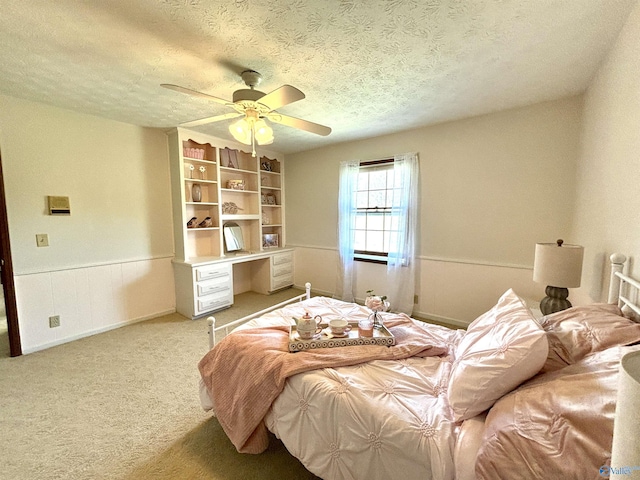 bedroom with a wainscoted wall, a textured ceiling, a ceiling fan, and light carpet