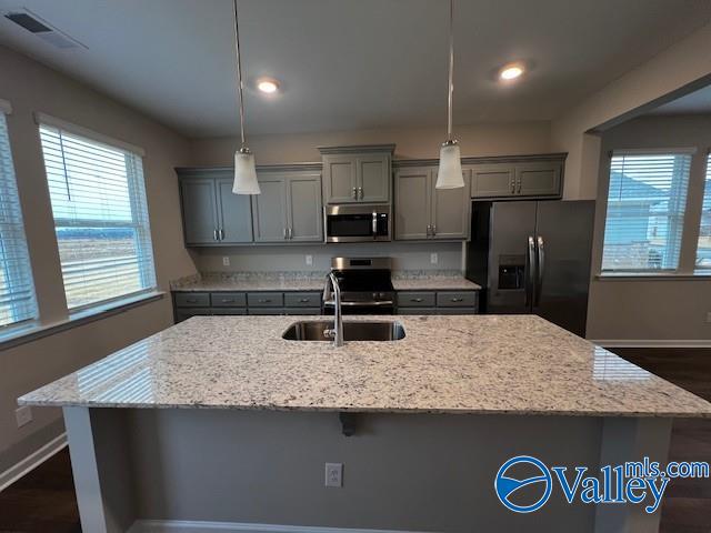 kitchen with dark wood-type flooring, hanging light fixtures, light stone countertops, an island with sink, and stainless steel appliances