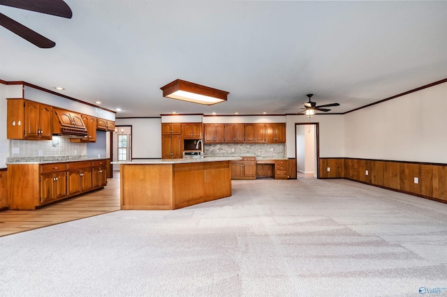 kitchen with light colored carpet, a center island with sink, ornamental molding, ceiling fan, and backsplash