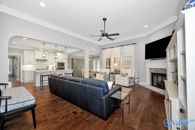 living room with arched walkways, a stone fireplace, ornamental molding, and ceiling fan