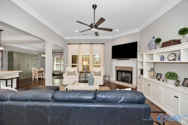 living room with a stone fireplace, ceiling fan with notable chandelier, ornamental molding, dark wood-style flooring, and recessed lighting