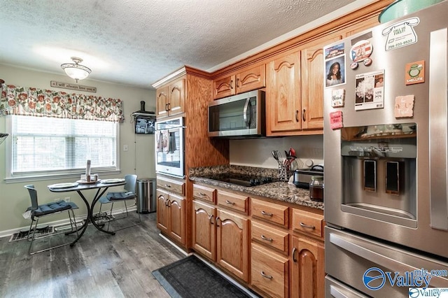 kitchen featuring dark wood-type flooring, a textured ceiling, dark stone counters, appliances with stainless steel finishes, and brown cabinetry