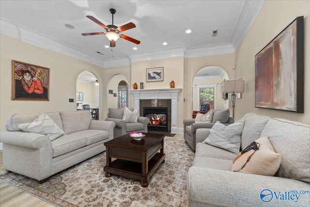 living room featuring arched walkways, ceiling fan, a premium fireplace, ornamental molding, and light wood-type flooring