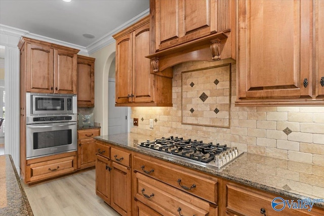 kitchen with stainless steel appliances, backsplash, dark stone counters, brown cabinetry, and crown molding