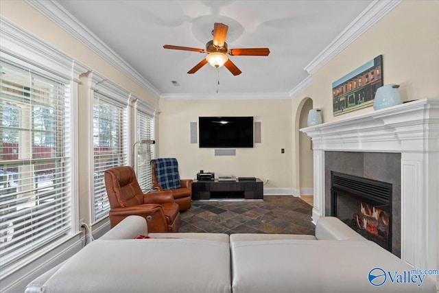 living room with arched walkways, a fireplace, ornamental molding, ceiling fan, and baseboards