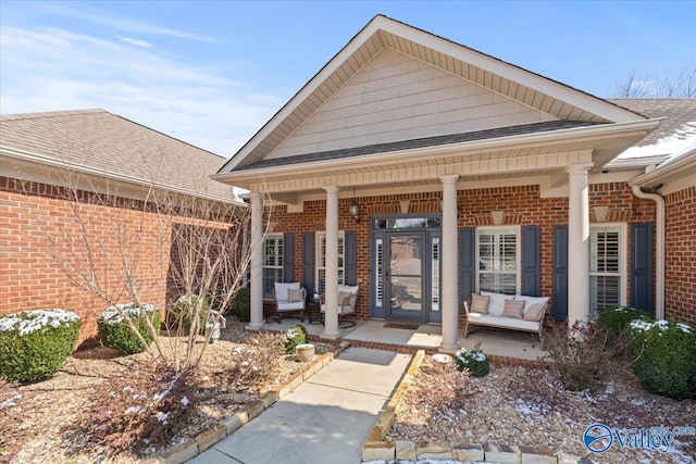 view of exterior entry with covered porch, roof with shingles, and brick siding