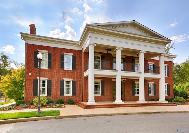 greek revival house with a balcony, a chimney, a ceiling fan, and brick siding