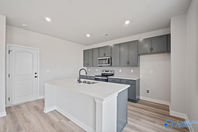 kitchen featuring a kitchen island with sink, gray cabinetry, a sink, light wood-style floors, and appliances with stainless steel finishes