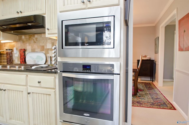 kitchen with ornamental molding, under cabinet range hood, backsplash, dark countertops, and appliances with stainless steel finishes