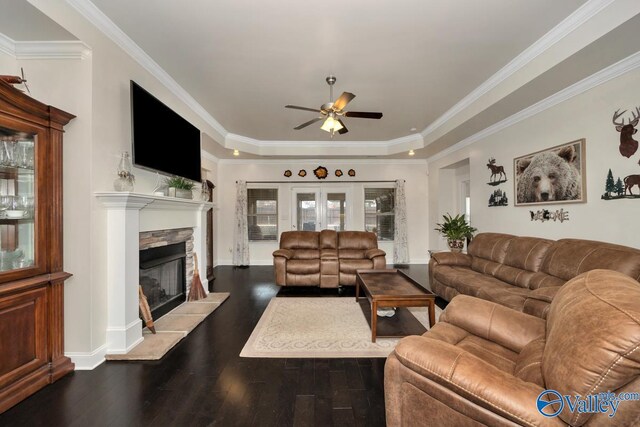 living room featuring a stone fireplace, ornamental molding, ceiling fan, a raised ceiling, and dark wood-type flooring