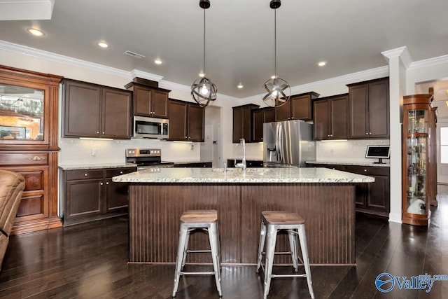 kitchen featuring stainless steel appliances, an island with sink, hanging light fixtures, and light stone countertops