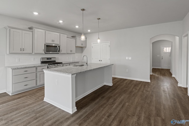 kitchen featuring arched walkways, a sink, appliances with stainless steel finishes, an island with sink, and dark wood finished floors