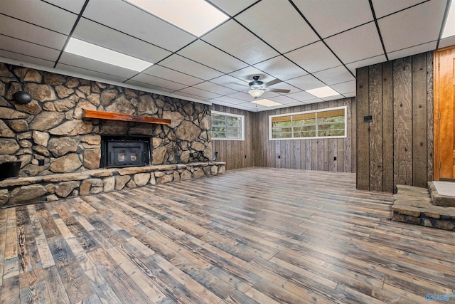 unfurnished living room featuring a wood stove, ceiling fan, a drop ceiling, wood walls, and hardwood / wood-style flooring