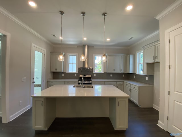 kitchen with wall chimney exhaust hood, sink, crown molding, hanging light fixtures, and a center island with sink