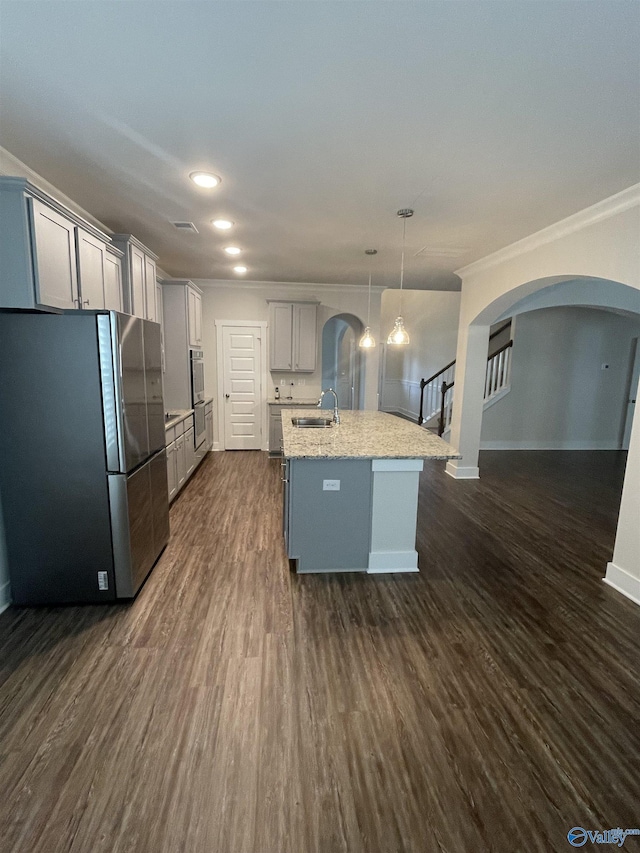 kitchen with light stone counters, a kitchen island with sink, dark wood-type flooring, stainless steel refrigerator, and hanging light fixtures