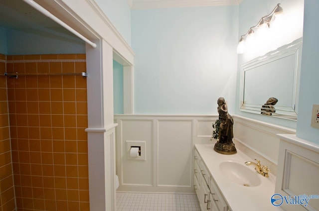 bathroom featuring tile patterned flooring, a decorative wall, vanity, tiled shower, and wainscoting