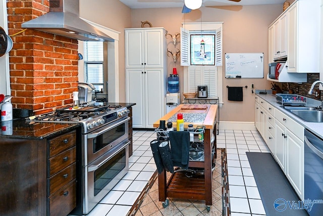 kitchen featuring light tile patterned flooring, ceiling fan, a sink, appliances with stainless steel finishes, and wall chimney range hood