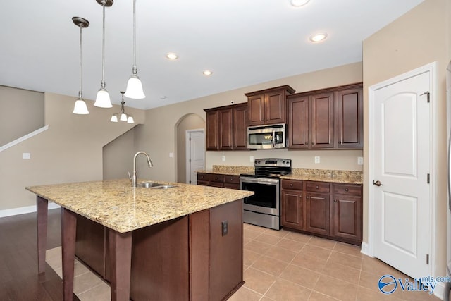 kitchen with light stone counters, an island with sink, arched walkways, a sink, and stainless steel appliances