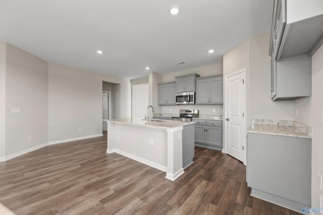 kitchen featuring dark wood-type flooring, stainless steel appliances, a center island with sink, and gray cabinetry