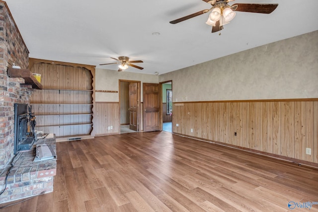 unfurnished living room featuring a brick fireplace, light hardwood / wood-style floors, wooden walls, and ceiling fan