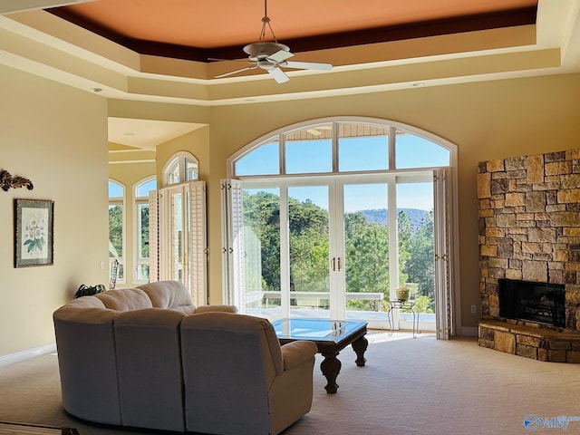 carpeted living room with a tray ceiling, a stone fireplace, and ceiling fan