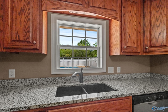 kitchen with brown cabinetry, light stone countertops, stainless steel dishwasher, and a sink