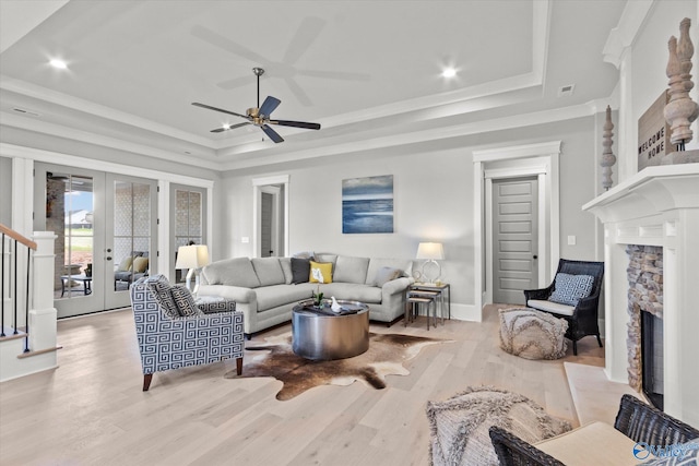 living room featuring a stone fireplace, french doors, light wood-type flooring, ceiling fan, and a raised ceiling