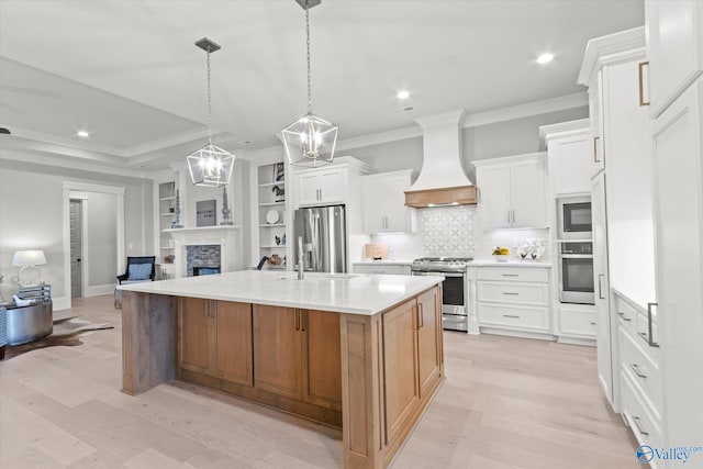 kitchen with white cabinetry, custom exhaust hood, stainless steel appliances, a stone fireplace, and a kitchen island with sink