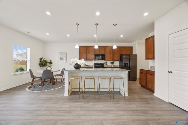 kitchen with light hardwood / wood-style flooring, decorative light fixtures, light stone countertops, black appliances, and a center island with sink