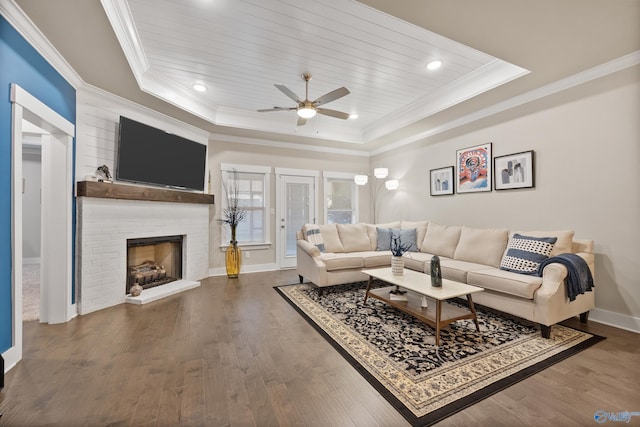 living room featuring dark wood-type flooring, a tray ceiling, crown molding, a fireplace, and ceiling fan