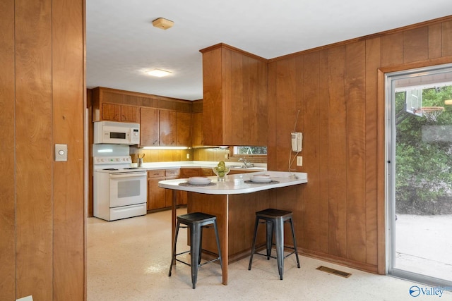 kitchen with light countertops, brown cabinetry, white appliances, a peninsula, and a kitchen breakfast bar