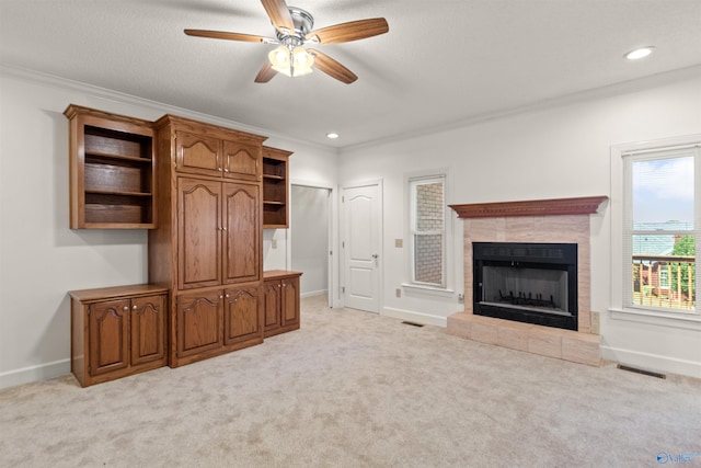 unfurnished living room with a textured ceiling, a tiled fireplace, light colored carpet, crown molding, and ceiling fan