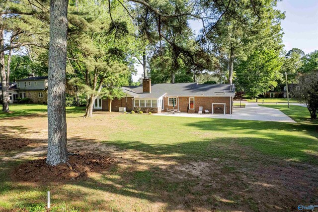 back of house featuring a chimney, brick siding, a patio, a lawn, and view of wooded area