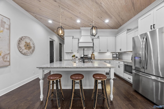 kitchen featuring premium range hood, a kitchen island with sink, stainless steel appliances, light stone countertops, and decorative light fixtures