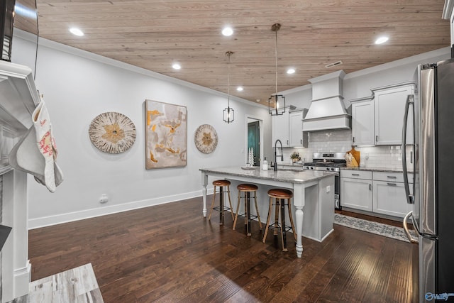 kitchen with pendant lighting, premium range hood, a kitchen island with sink, appliances with stainless steel finishes, and light stone counters