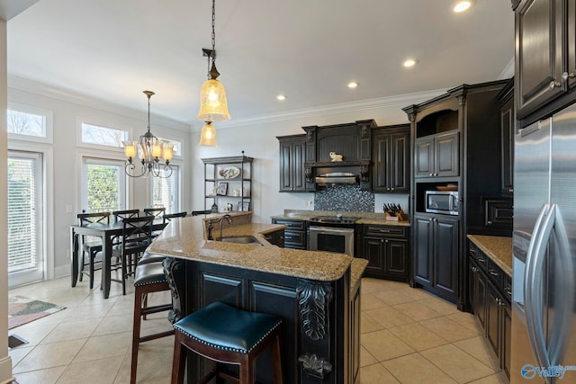 kitchen with light stone counters, an island with sink, stainless steel appliances, decorative light fixtures, and sink