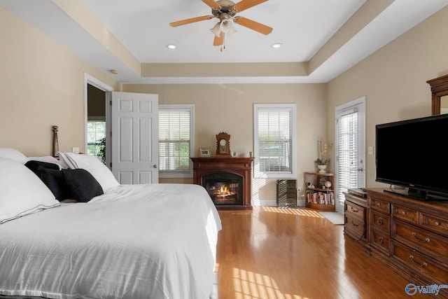 bedroom with light wood finished floors, a glass covered fireplace, a tray ceiling, and multiple windows