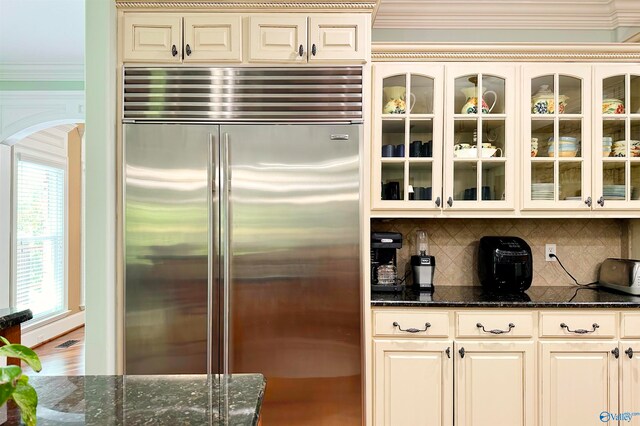 kitchen with dark stone counters, stainless steel built in fridge, decorative backsplash, cream cabinetry, and ornamental molding