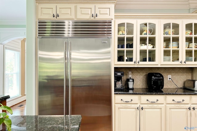 kitchen featuring dark stone counters, built in fridge, and glass insert cabinets