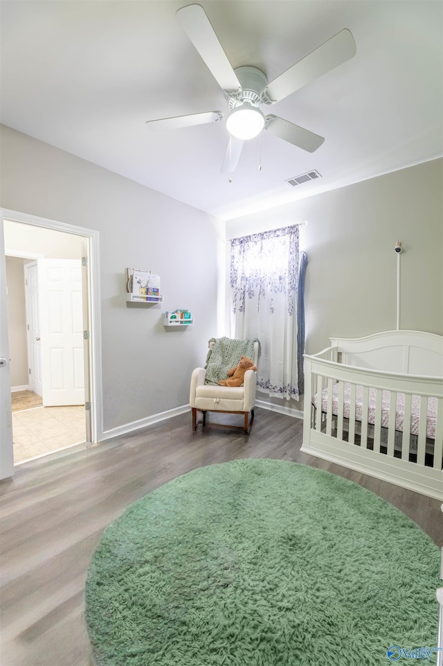 bedroom with visible vents, baseboards, a nursery area, wood finished floors, and a ceiling fan