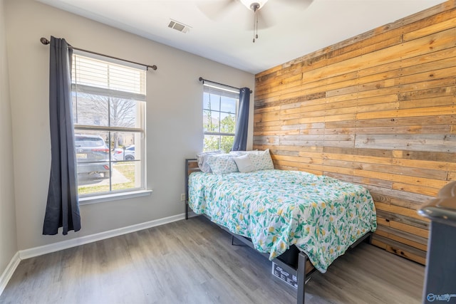 bedroom with wooden walls, wood finished floors, visible vents, baseboards, and ceiling fan