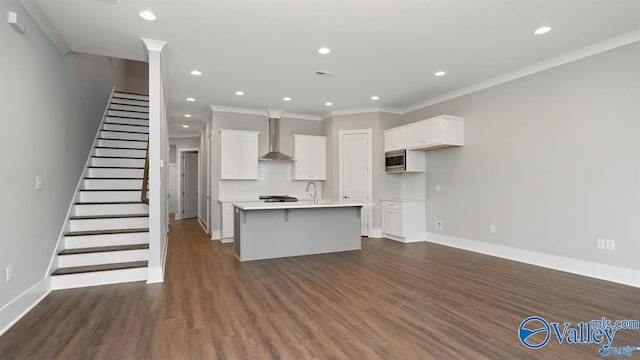 kitchen with crown molding, a kitchen island with sink, stainless steel microwave, white cabinets, and wall chimney exhaust hood