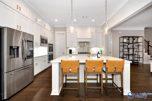 kitchen with decorative light fixtures, custom range hood, stainless steel appliances, a kitchen island with sink, and white cabinets